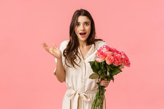 Surprised and happy, excited pretty young woman receive flowers and looking astonished by romantic nice gesture, didnt expact feeling touched raise hand smiling, hold roses, pink background.
