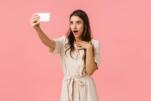 Girl making fascinated and surprised expression as talking to girlfriend on phone video-call. Charming brunette female in dress, taking selfie, gasping open mouth amused, standing pink background.