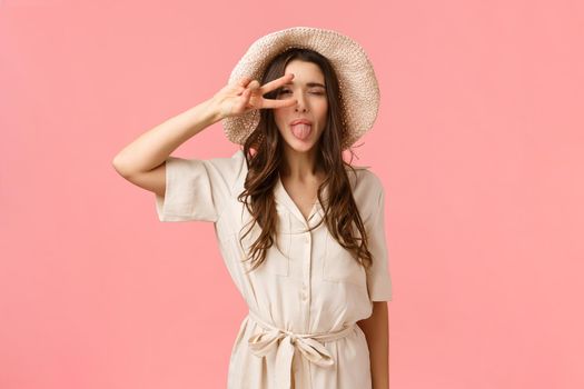 Cheerful and beautiful young brunette female enjoying lovely day, feeling ecstatic and overjoy, showing peace sign near eye, close eyes and stick tongue happily, standing pink background.