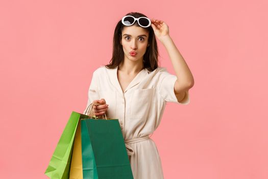 Glamour feminine caucasian girl having fun relaxing, holding shopping bags and folding lips amused, taking-off glasses seeing cool product, want buy, making purchase, standing pink background.