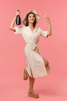 Full-length vertical portrait happy charismatic young woman in hat and dress, dancing with raised bottle champagne and dress, laughing enjoying celebrating occasion, standing pink background.