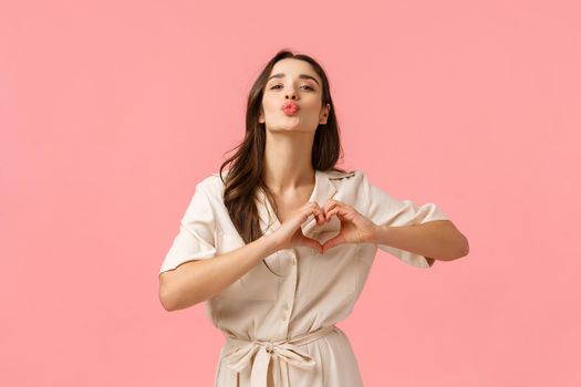 Fashion, beauty and tenderness concept. Charming gorgeous brunette female in cute dress, showing heart love gesture, folding lips for kiss, express romantic feelings, affection, pink background.