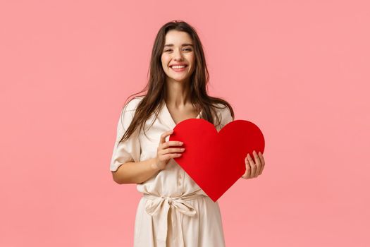 Silly and carefree, lovely alluring brunette girl in fancy dress, holding red big heart card, having best valentines day ever, smiling and laughing delighted, standing pink background upbeat.