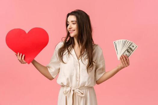 Waist-up portrait surprised and determined brunette woman making her choice towards true love, looking at heart, holding valentines card and cash, dont care wealth and money, pink background.