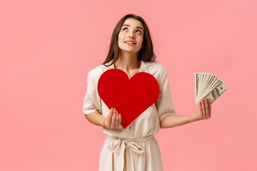 Dreamy young and cute girl imaging things, wanting find true love dont care money. Attractive alluring woman looking up thoughtful and smiling, holding heart card and cash dollars, pink background.