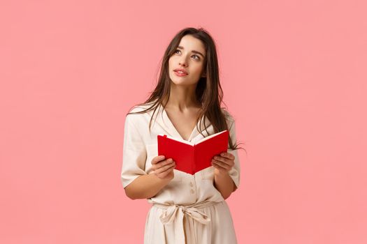 Thoughtful and inspired, creative female student having inspiration, looking up dreamy and wondered, holding red notebook, learning or preparing to class, standing pink background.