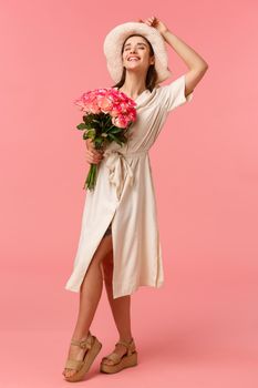 Full-length vertical portrait gorgeous stylish young european woman in dress and hat, close eyes and smiling as enjoying warm sunlights on her face, holding bouquet roses, pink background.