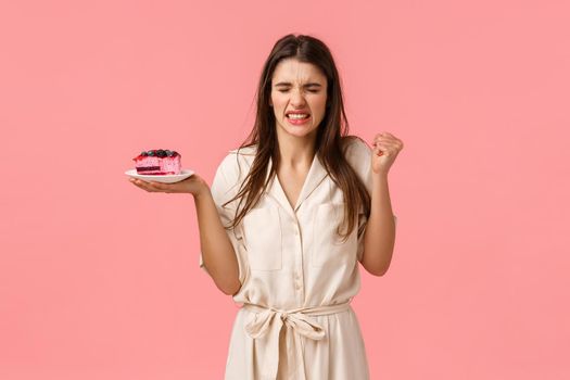 Girl gonna puke from disgusting taste of cake. Disappointed young woman try eating dessert and grimacing, squinting from awful smell, holding dessert and standing pink background.