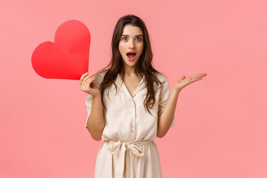 Surprise, relationship and love concept. Amused and wondered alluring woman didnt expact receive valentines day confession, open mouth gasping excited, holding red heart, pink background.
