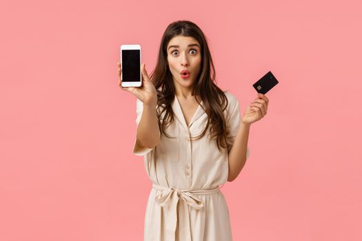 Girlfriend wasted all money on bank account during shopping discount event, holding credit card folding lips silly, showing smartphone screen, standing amazed pink background. Copy space