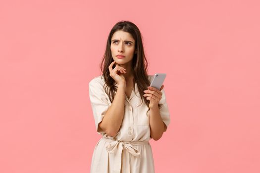 Troubled young girl like shopping, cant decide what order, holding smartphone turn left thoughtful and focused, frowning squinting pensive, making decision, standing pink background unsure.