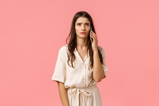 Serious and stressed young brunette woman hearing something strange, waiting someone pick-up phone, standing over pink background in dress, holding smartphone near ear, pink background.
