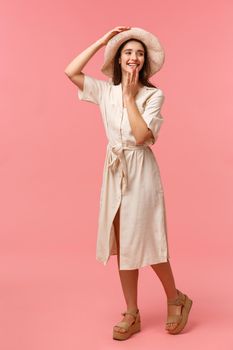 Full-length vertical portrait gorgeous, feminine lovely girl having fun, walking and enjoying sunny day, wearing hat and dress, amused looking right and cover mouth, pink background.