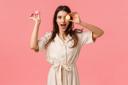 Excitement, holidays and sweet desserts concept. Elegant attractive young woman enjoy eating delicious food, holding macarons on on eye, open mouth gasping amused, standing pink background.