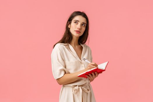 Inspiration, creativity and education concept. Concentrated pensive and creative young girl using imagination, thinking looking up wondered stop for second as writing something in red notebook.