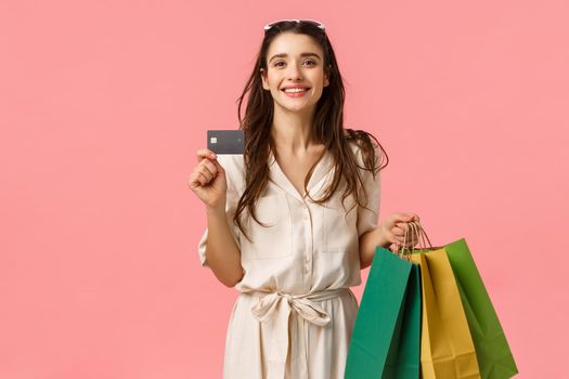 Happy caucasian brunette woman in stylish dress, carry shopping bags and credit card, paying everything with paypass, have special discount, using bank deposit, standing pink background.