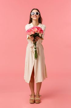 Full-length vertical portrait attractive sassy and stylish brunette woman giving flowers to her girlfriend, extending hands towards camera holding nice bouquet roses, standing pink background.