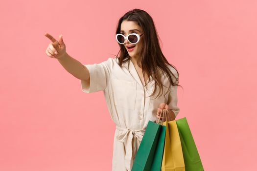 Sassy and elegant young woman in dress, seeing exactly what need, holding shopping bags, pointing finger sideways and looking determined camera, standing pink background excited.