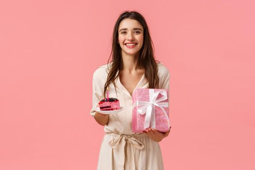 B-day girl feelings happy and upbeat, enjoying celebrating. Attractive caucasian female receive presents, holding delicious piece cake, wrapped present, smiling joyfully, standing pink background.