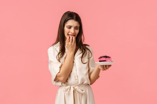 Cant resist temptation. Young woman on diet, trying eat healthy food want take bite of delicious blueberry cake, biting fingernails and staring on plate, standing pink background with desire.