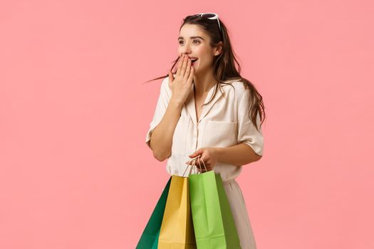 Excited and fascinated cute feminine brunette female in dress, holding shopping bags, wonder around mall, gasping excited cover mouth seeing something wonderful, pink background. Copy space