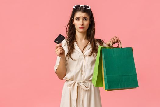 Upset and distressed young brunette female feeling sad spent all money, looking uneasy and concerned at credit card, holding shopping bags, standing pink background gloomy. Copy space