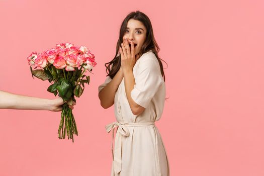 Valentines day, delivery and beauty concept. Amazed woman gasping and looking camera surprised as someone giving her beautiful bouquet flowers, congratulations with anniversary, pink background.