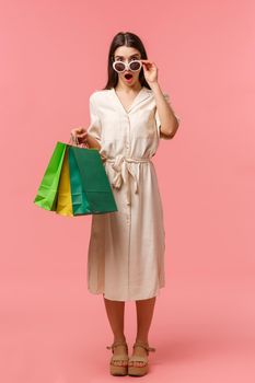 Full-length vertical portrait excited and speechless, amazed young brunette woman seeing something amazing and pretty, taking off glasses to look closer, open mouth fascinated, holding shopping bags.