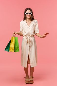 Full-length vertical portrait elegant good-looking woman on vacation, shopping and having fun in malls, wearing sunglasses, light dress, carry bags from store, standing pink background smiling.