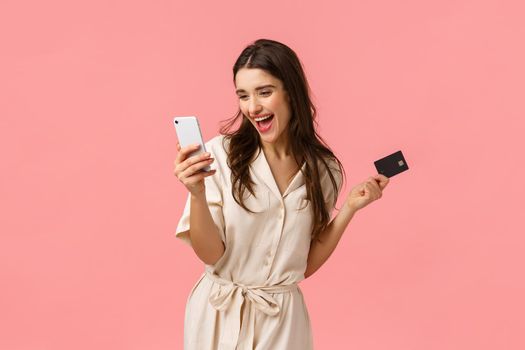 Excitement, shopping and holidays concept. Cheerful and carefree smiling pretty woman making online purchase, paying for internet delivery, cant wait receive product, hold credit card and smartphone.