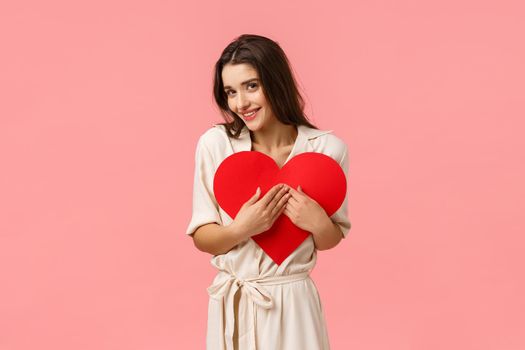 Tenderness, romance and care concept. Attractive silly girl in dress, embracing heart card, holding valentines sign and coquettish glancing camera, smiling delighted, standing pink background.