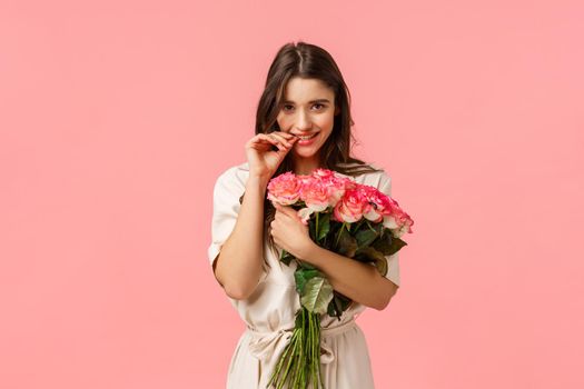 Romantic date, beauty and fashion concept. Gorgeous brunette girl in dress, receive beautiful bouquet flowers, got roses, biting finger and gazing camera flirty, standing pink background.