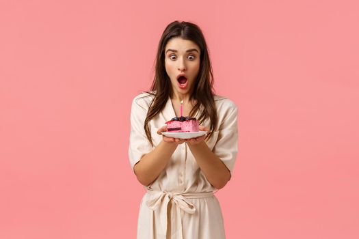 Celebration, birthday and holidays concept. Excited and surprised female in dress, received delicious piece cake blowing-out candle and making wish, gasping amused, standing pink background.