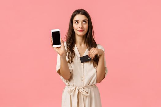Dreamy and coquettish good-looking young woman in dress, dreaming waiting for delivery order online, holding smartphone and credit card, showing mobile screen, standing pink background.