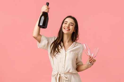 Party, holidays and celebration concept. Cheerful lucky young brunette girl having fun, dancing raising bottle champagne and glass, close eyes and smiling carefree, standing pink background.