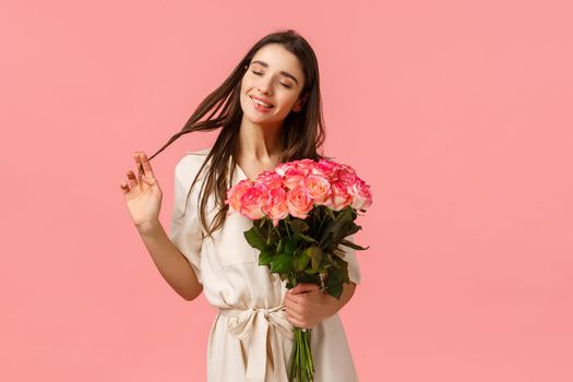 Tenderness, delight and valentines day concept. Charming, lovely and sensual brunette woman in dress, rolling curl on hair happily close eyes and dreaming, receive flower delivery, hold roses.