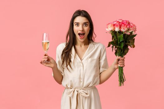 Wonderful news, celebration and emotions concept. Cheerful attractive brunette female in dress, spread hands sideways surprised and wondered, open mouth gasping, hold champagne glass and flowers.