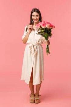 Full-length vertical portrait sensual, romantic and coquettish young woman seeing something interesting, looking with temptation or desire, holding bouquet flowers, received roses, pink background.