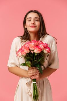 Full-length vertical portrait romantic and dreamy, sensual brunette girl in dress, holding beautiful bouquet flowers, smelling roses with closed eyes and smiling, standing happy over pink background.