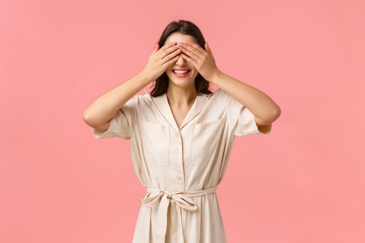 Romantic moment, beauty and tenderness concept. Attractive brunette girlfriend asking close eyes, cover sight with palms as waiting surprise gift, standing pink background, smiling with temptation.