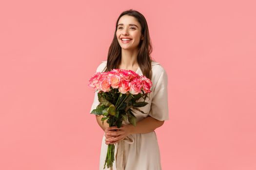 Valentines day, holidays, celebration and beauty concept. Charismatic attractive brunette female in dress, holding bouquet flowers, smiling delighted, receive present, standing pink background.