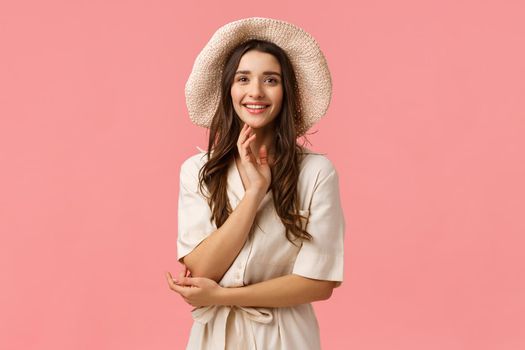Vacation, beauty and shopping concept. Cheerful tender glamour brunette female in dress, fancy hat, standing elegant and cute over pink background, smiling having casual conversation.