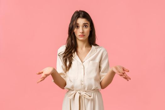 So what, I dont know. Silly and clueless uncertain cute, glamour girl in dress, shrugging and raise hands sideways confused, cant figure out what do, standing pink background unsure.