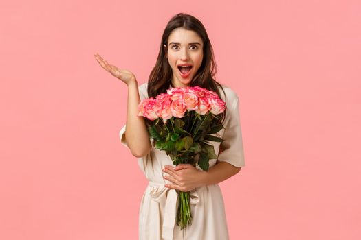 Wow such wonferful unexpected surprise. Charming lovely and romantic cute brunette girl in dress over pink wall, receive beautiful flowers, holding roses and looking amused, raise hand astonished.