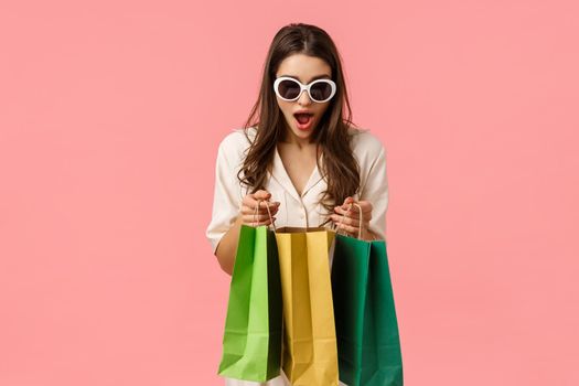 Shoppaholic, consumer and women concept. Excited and happy young attractive caucasian woman in glasses, holding shopping bags and looking inside amazed, standing pink background. Copy space