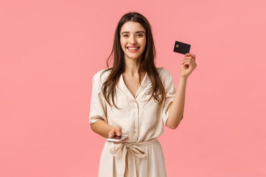 Advertising, technology and digital lifestyle concept. Carefree attractive young woman in gorgeous dress showing credit card and holding smartphone, smiling buying online, pink background.