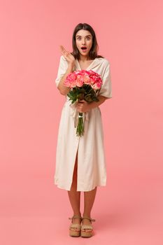 Full-length vertical portrait pretty surprised girl receive unexpected gift, got delivery holding flowers, gasping open mouth amazed and looking speechless with stunned expression, pink background.