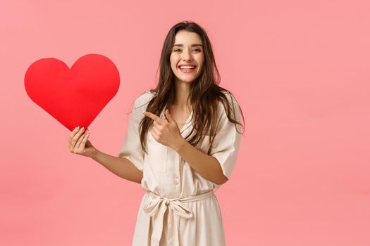 Searching for soulmate. Cheerful pretty laughing smiling girl in dress, holding big red heart, pointing valentines cardboard and gazing camera, want show affection and love, pink background.