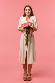 Full-length vertical portrait of dreamy, romantic woman in dress, receiving beautiful roses from secret admirer on birthday, close eyes and sniffing smell of flowers, standing pink background.