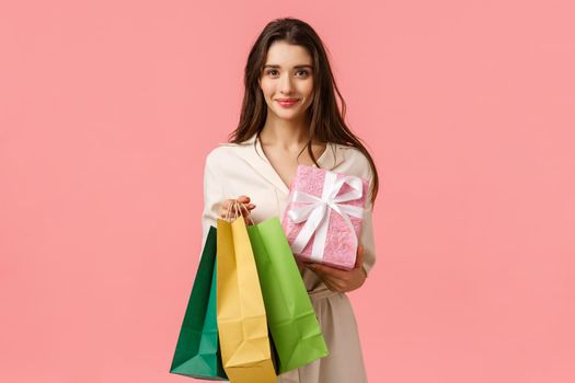 Cheerful feminine cute brunette woman holding shopping bags and wrapped gift, smiling, prepared surprise present for girlfriend, standing in dress over pink background happy. Copy space
